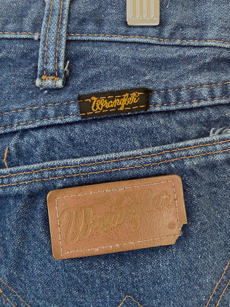 May include: Close-up of blue denim jeans with the brand name "Wrangler" visible on two separate tags. One tag is a small black rectangle with yellow stitching, and the other is a brown leather patch.