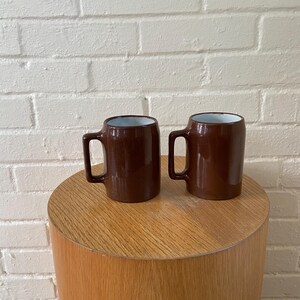 May include: Two brown ceramic mugs with handles, each with a white interior, sit on a light brown, cylindrical wooden table. The mugs have a glossy finish and a simple, classic design, perfect for coffee or tea.