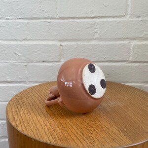 May include: A small, round, pink ceramic pitcher with a handle, resting upside down on a wooden table. The bottom of the pitcher has three black dots on a white surface. The table is a light brown color, and the background is a white brick wall.