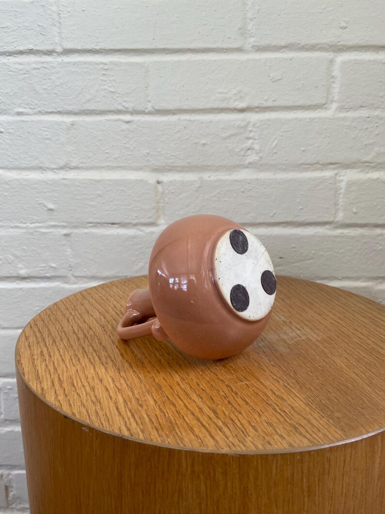 May include: A small, round, pink ceramic pitcher with a handle, resting upside down on a wooden table. The bottom of the pitcher has three black dots on a white surface. The table is a light brown color, and the background is a white brick wall.