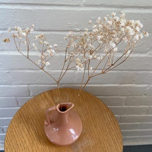 May include: A small, pink ceramic vase with a handle holds dried white flowers. The vase sits on a round, light-colored wooden table. The background is a white brick wall.