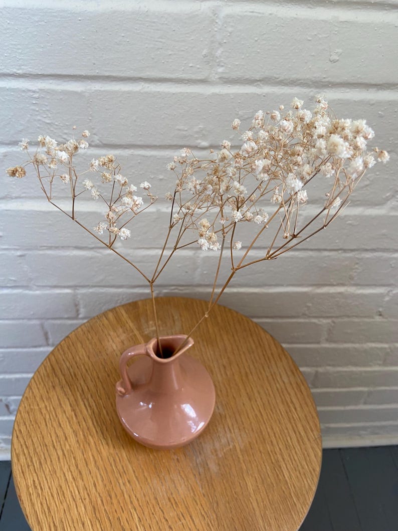May include: A small, pink ceramic vase with a handle holds dried white flowers. The vase sits on a round, light-colored wooden table. The background is a white brick wall.