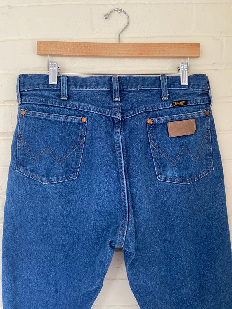 May include: Blue denim jeans with copper rivets and a brown leather patch. The jeans are hanging on a wooden hanger. The Wrangler logo is visible on the back pocket.