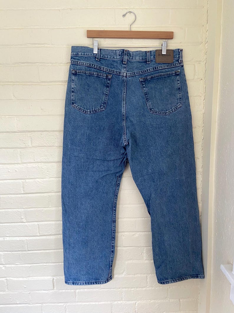 May include: Light blue denim jeans hanging on a wooden hanger. The jeans have two back pockets and a brown leather patch. The jeans are a classic, straight-leg style.