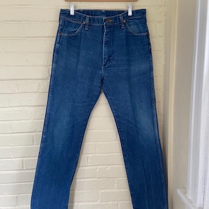 May include: Blue denim jeans hanging on a wooden hanger against a white brick wall. The jeans have a classic five-pocket design, a button and zipper closure, and a straight leg fit. The denim has a medium wash with some fading.
