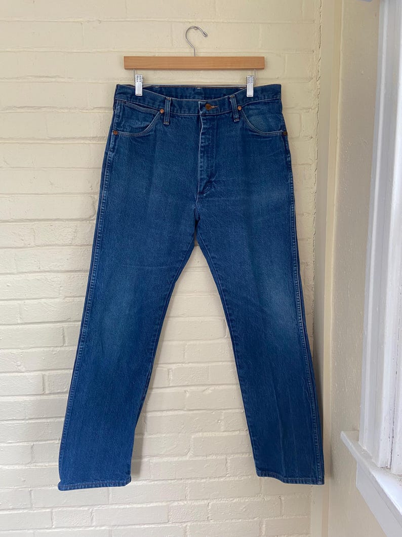 May include: Blue denim jeans hanging on a wooden hanger against a white brick wall. The jeans have a classic five-pocket design, a button and zipper closure, and a straight leg fit. The denim has a medium wash with some fading.