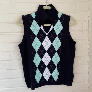 May include: A sleeveless navy blue sweater vest with a high collar and a keyhole detail. The front features a diamond pattern in mint green and white. The vest is hanging on a hanger against a white brick wall.