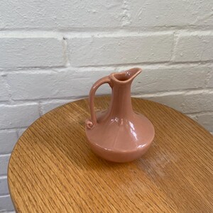 May include: A small, pink ceramic pitcher with a handle and spout. The pitcher has a rounded body and sits on a wooden table. The background is a white brick wall. The pitcher is likely vintage or antique.