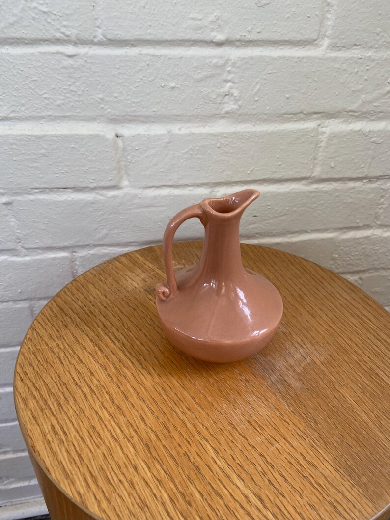 May include: A small, pink ceramic pitcher with a handle and spout. The pitcher has a rounded body and sits on a wooden table. The background is a white brick wall. The pitcher is likely vintage or antique.