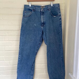May include: Light blue denim jeans hanging on a wooden hanger. The jeans have a classic five-pocket design, a button and zipper closure, and a straight leg fit. The jeans are made of a durable denim fabric and are suitable for everyday wear.