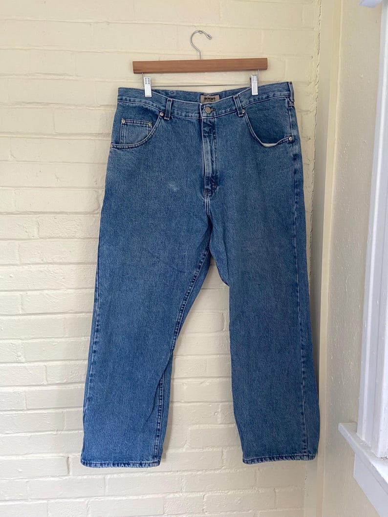 May include: Light blue denim jeans hanging on a wooden hanger. The jeans have a classic five-pocket design, a button and zipper closure, and a straight leg fit. The jeans are made of a durable denim fabric and are suitable for everyday wear.