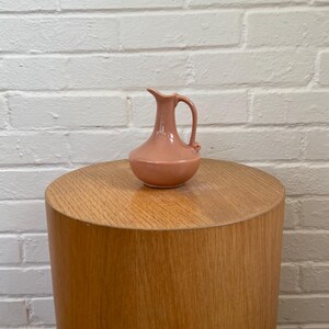 May include: A small, peach-colored ceramic pitcher with a handle and spout, resting on a cylindrical wooden side table. The table has a light wood grain finish. The background is a white brick wall.
