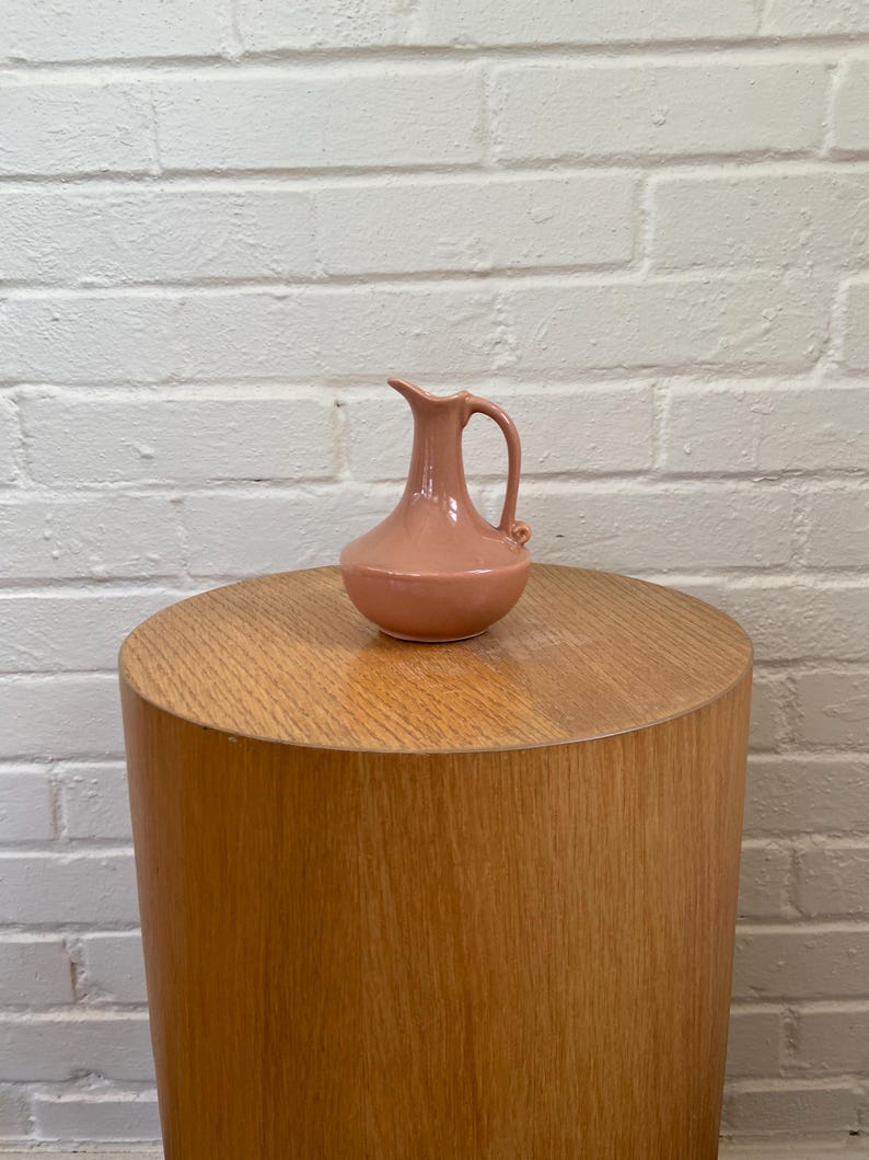 May include: A small, peach-colored ceramic pitcher with a handle and spout, resting on a cylindrical wooden side table. The table has a light wood grain finish. The background is a white brick wall.