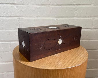 May include: A rectangular, dark brown wooden box with inlaid mother-of-pearl diamond and circular accents. The box is displayed on a light brown, cylindrical wooden stand against a white brick wall.