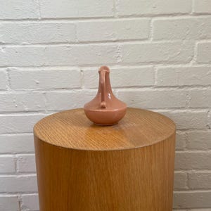 May include: A small, peach-colored ceramic pitcher with a handle and spout. The pitcher is set on a cylindrical wooden side table. The background is a white brick wall.