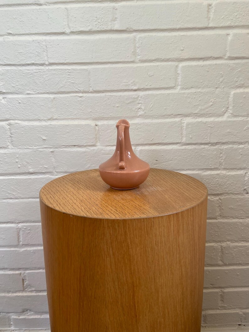 May include: A small, peach-colored ceramic pitcher with a handle and spout. The pitcher is set on a cylindrical wooden side table. The background is a white brick wall.