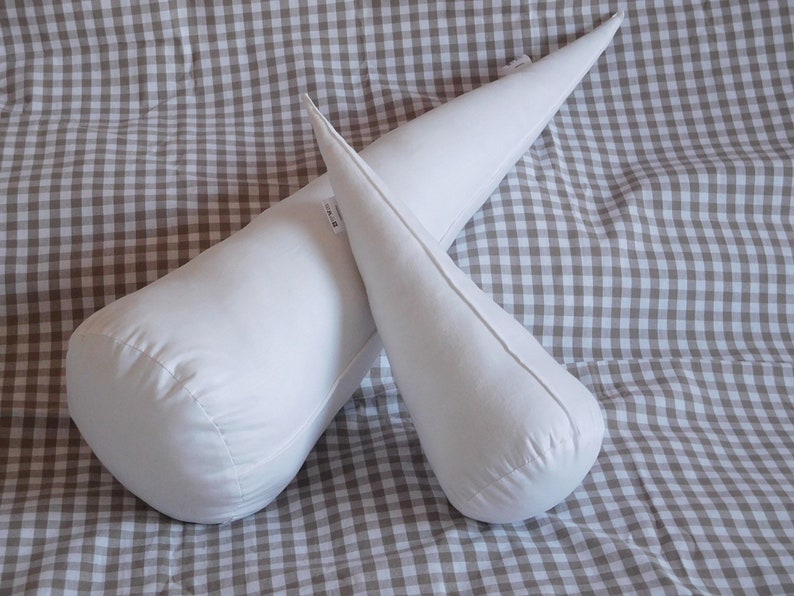 May include: Two white fabric pillows, one shaped like a cone and the other shaped like a long cylinder, are lying on a brown and white checkered fabric.