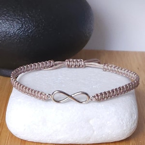 Può includere: Un braccialetto in corda intrecciata marrone chiaro con un simbolo dell'infinito color argento al centro. Il braccialetto è dotato di un meccanismo di chiusura a nodo scorsoio regolabile ed è appoggiato su una pietra bianca liscia.