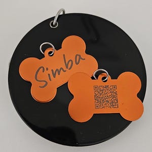 Könnte beinhalten: Zwei orangefarbene, knochenförmige Hundemarken. Eine Marke ist mit dem Namen "Simba" graviert, die andere Marke hat einen schwarz-weißen QR-Code.
