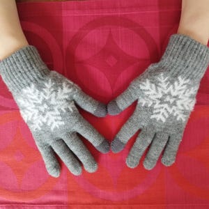 Könnte beinhalten: Graue Strickhandschuhe mit weißen Schneeflockenmustern. Die Handschuhe haben graue Fingerspitzen und Bündchen. Die Handschuhe werden auf einer roten Oberfläche gezeigt.