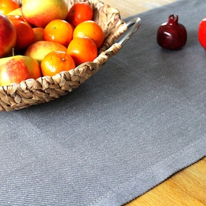 Könnte beinhalten: Ein geflochtener Korb mit Äpfeln und Mandarinen steht auf einem grauen Leinenläufer. Zwei dekorative Granatapfelskulpturen, eine dunkelrote und eine leuchtend rote, sind auf dem Läufer platziert.