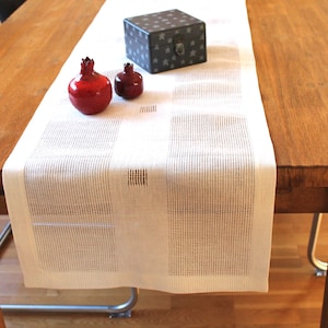 Könnte beinhalten: Ein Holztisch mit einem cremefarbenen Tischläufer. Dekorative Elemente sind zwei rote Granatapfel-Ornamente und eine kleine schwarze Box mit Sternmuster. Der Tisch ist von schwarzen Stühlen umgeben.