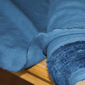 May include: A roll of indigo blue fabric, with a raw, unfinished edge. The fabric is tightly rolled, revealing layers of the material. The texture appears soft and slightly wrinkled, suggesting a natural fiber like linen or cotton.