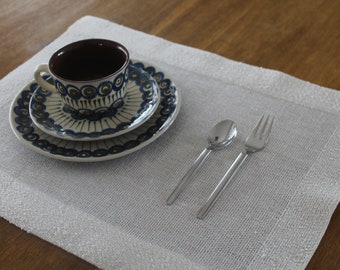 May include: A ceramic teacup and saucer set with a blue and white floral design, along with a matching plate. A silver spoon and fork rest on a white placemat. The items are arranged on a wooden surface.