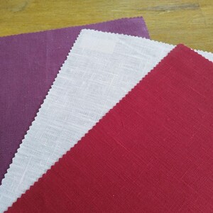 May include: Three fabric swatches in shades of purple, white, and red. The fabrics have a textured appearance and are arranged diagonally, showcasing their colors and textures. The edges are cut with a zig-zag pattern.