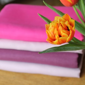 May include: A stack of four fabric swatches in shades of pink, white, and purple. A bouquet of orange and pink tulips with green leaves is in front of the fabric.