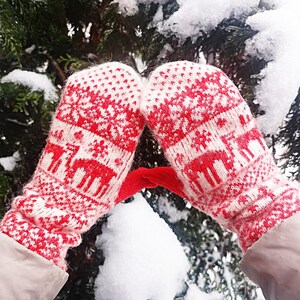 Könnte beinhalten: Ein Paar Strickfäustlinge mit einem rot-weißen Fair-Isle-Muster, das Rentier- und Schneeflockenmotive zeigt. Die Fäustlinge haben ein geripptes Bündchen und eine strukturierte Strickoptik mit einem durchgehenden geometrischen Muster.