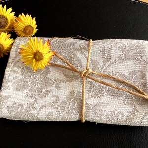 Könnte beinhalten: Ein rechteckiges, gefaltetes Textil mit einem hellbeigen und weißen floralen Jacquardmuster. Der Stoff ist mit einer dünnen, naturfarbenen Schnur zusammengebunden, die einen einfachen Knoten bildet. Drei leuchtend gelbe Strohblumen liegen obenauf.