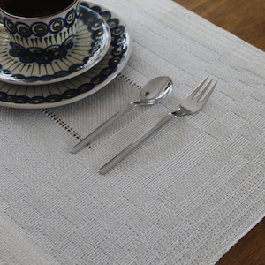 Puede incluir: Un mantel individual tejido blanco con una taza de té y un platillo con un patrón azul y blanco, un tenedor de plata y una cuchara de plata.