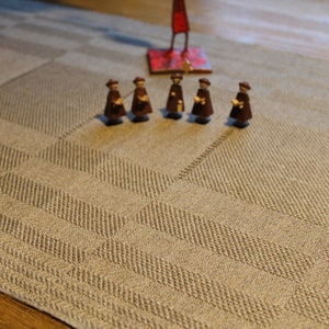 Könnte beinhalten: Eine Gruppe von sieben kleinen Holzfiguren, die braune Roben tragen, stehen auf einem beigefarbenen Tischläufer mit einem karierten Muster. Die Figuren sind in einer Reihe angeordnet und nach vorne gerichtet.