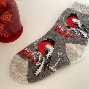 May include: A grey knit sock featuring a repeating pattern of bullfinch birds with red chests and black wings perched on branches with red berries. The sock has a light grey ribbed cuff, a light grey heel, and a light grey toe section.
