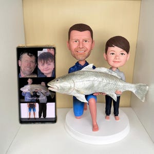May include: A custom bobblehead figurine featuring an adult and a child holding a large silver fish, standing on a round white base. Next to the figurine is a smartphone displaying a photo collage of the same pair with the text Fresh bait pays off!