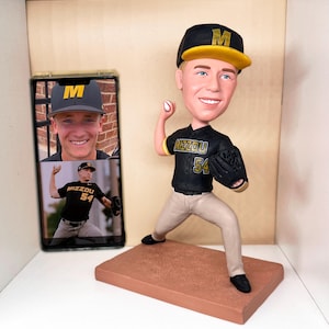 Könnte beinhalten: Eine Bobblehead-Figur eines Baseball-Pitchers in einer schwarz-goldenen Uniform mit dem Wort "Mizzou" und der Nummer "54". Die Figur steht auf einer braunen Basis und trägt eine schwarz-goldene Baseballkappe. Ein Smartphone zeigt zwei Fotos einer Person mit derselben Kappe und Uniform.