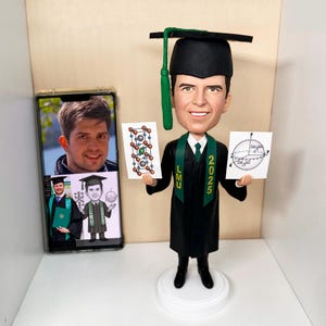 May include: A graduation bobblehead of a smiling person in a black cap and gown, holding two cards with diagrams. The bobblehead has a green tassel and a green sash with "LMU 2025". A smartphone displays a photo of the person, with a similar graduation theme.