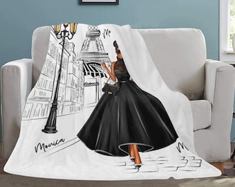 May include: White throw blanket featuring a black and white illustration of a woman in a black dress in Paris. The Eiffel Tower and a street scene are in the background. The name "Monica" is written on the blanket.