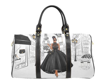 May include: A white travel bag featuring a black and white illustration of a woman in a black dress in Paris. The bag has black leather handles and trim, with gold-colored hardware. The Eiffel Tower and a cafe are visible in the background.