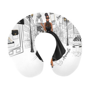 May include: A white travel neck pillow with a black and white illustration of a woman in a black dress walking in Paris. The Eiffel Tower is in the background. The text "Amanda" is written on the pillow.