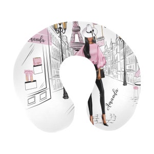 May include: A white travel neck pillow with a black and white illustration of a woman in a pink top and black pants walking in a Parisian street scene. The text "Amanda" appears twice on the pillow.