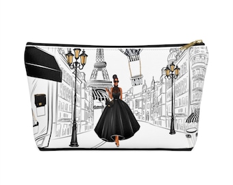 May include: A white makeup bag with a black and white illustration of a woman in a black dress walking down a street in Paris. The Eiffel Tower is in the background.