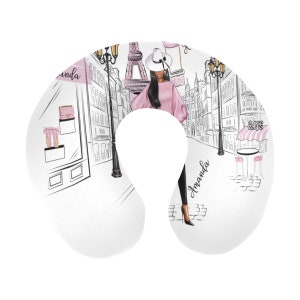 May include: A white travel pillow with a pink and black illustration of a woman in a pink sweater and black pants walking in Paris. The Eiffel Tower is in the background. The text "Amanda" is written on the pillow.