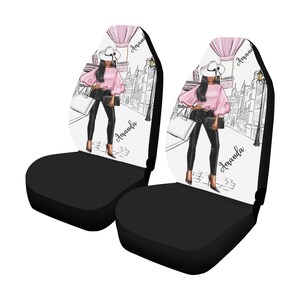 May include: A pair of black car seat covers with a white background featuring a stylized illustration of a woman in a pink top and black pants walking down a city street. The text "Amanda" is written on each cover.