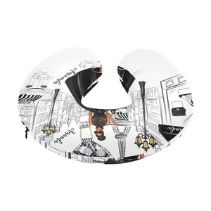 May include: A white travel neck pillow with a black and white illustration of a woman in a black dress walking in a Parisian street scene. The illustration includes the Eiffel Tower, street lamps, and the text "Journee".