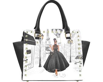 May include: Black and white handbag with a graphic print of a woman in a black dress walking in front of the Eiffel Tower. The handbag has a black leather handle and gold studs.
