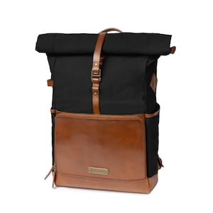 Könnte beinhalten: Schwarzer und brauner Rucksack mit Rollverschluss. Der Rucksack hat eine braune Lederfronttasche und Riemen mit einem schwarzen Canvas-Körper. Die Tasche hat Seitentaschen und einen Metall-Schnallenverschluss. Die Abmessungen betragen ca. 46 cm hoch, 30 cm breit und 15 cm tief.