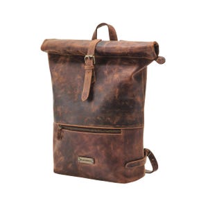 Puede incluir: Una mochila de cuero marrón con cierre enrollable, asa superior, correa vertical con hebilla de metal y un bolsillo frontal horizontal con cremallera. Una pequeña placa metálica rectangular con el texto DRAKENSBERG está fijada al frente.