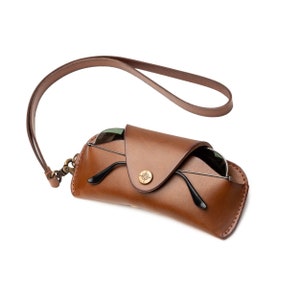 Puede incluir: Un estuche de gafas de cuero marrón con una correa. El estuche tiene una solapa que se cierra con un botón de latón. El estuche está vacío y la correa está suelta.